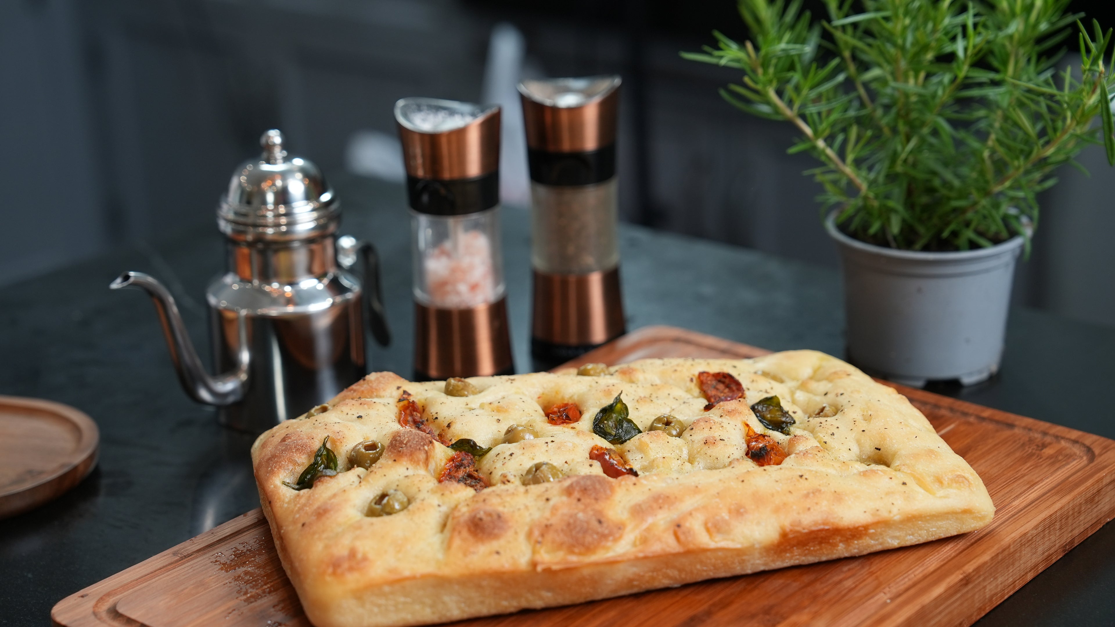 Loaf of focaccia on a wooden cutting board with a teapot, salt and pepper shakers, and a plant in the background.