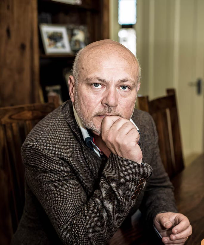 Giovanni in a brown suit sitting at a table with a serious expression.