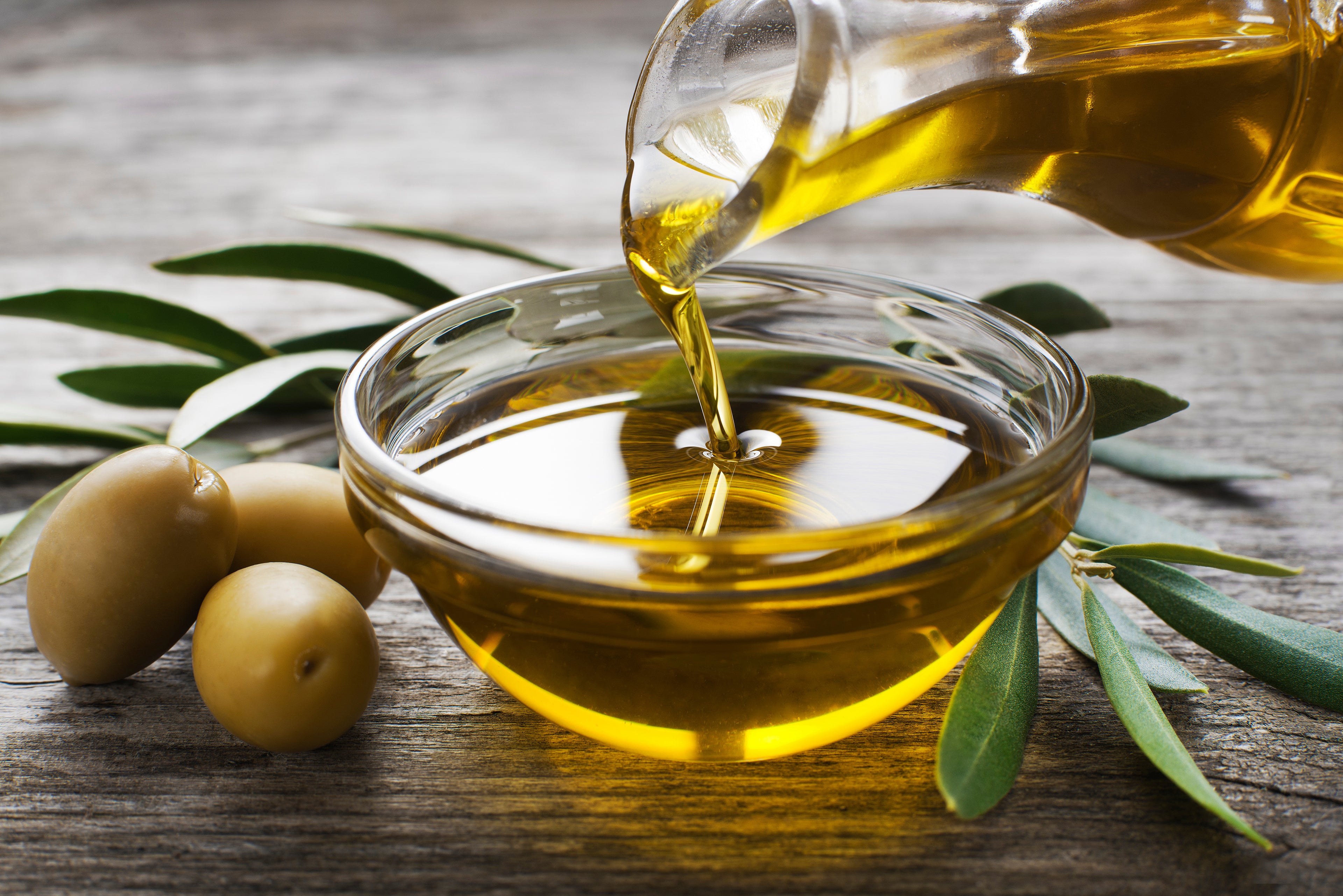 Olive oil being poured into a glass bowl with olives and olive leaves on a wooden surface